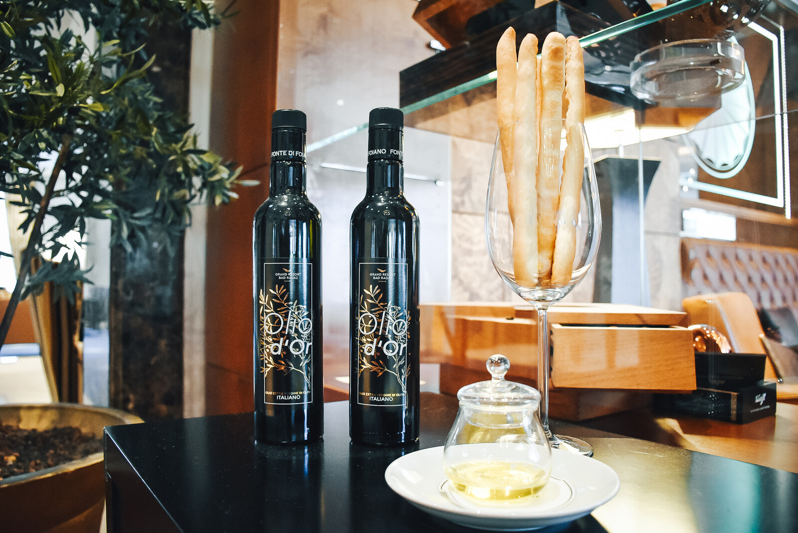 Zwei Ölflaschen stehen neben einem Weinglas mit Brotstecken. Neben diesem befindet sich ein kleines Glastöpfchen, in welchem sich Öl befindet. Auf den beiden Flaschen befindet sich jeweils ein Etikett auf dem "Olio d'Or" steht. Die Flaschen gehören zu dem Restaurant Olives d'Or, welches sich in dem Superior Hotel Grand Resort Bad Ragaz in der Schweiz befindet.