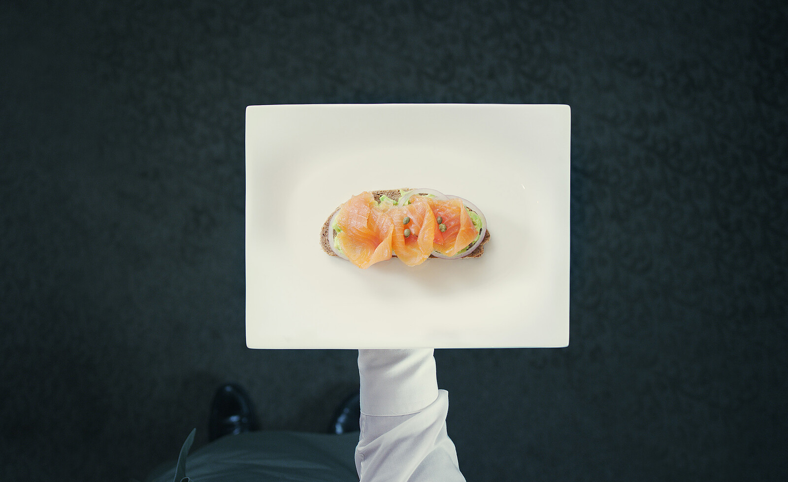 On a white plate lies a slice of bread topped with salmon and onions. A waiter is about to serve this. The Olives d'Or restaurant is located in the Hotel Grand Resort Bad Ragaz, a superior luxury resort in Switzerland.