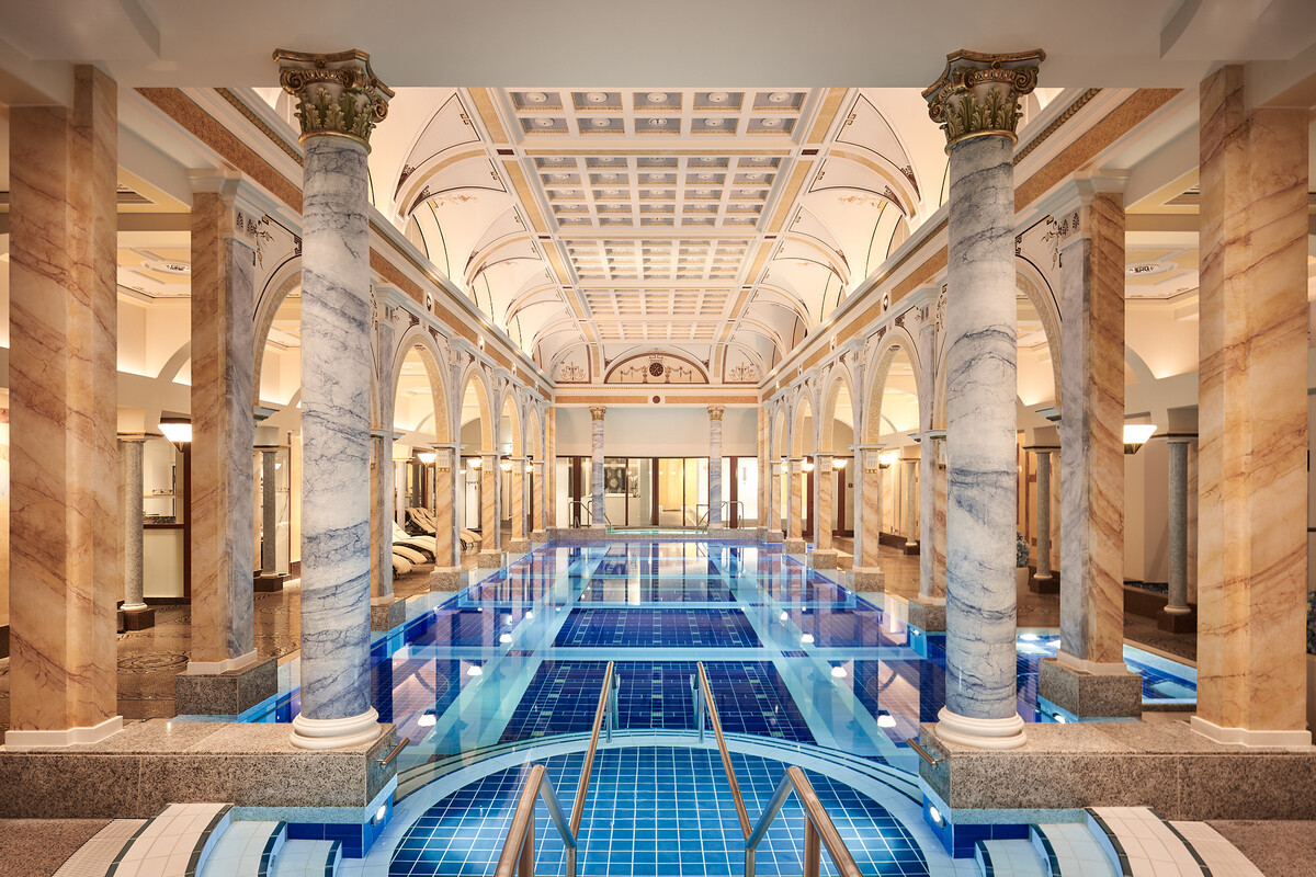 Luxuriöses Helenabad mit blauen Fliesen, umgeben von Marmorsäulen und Bögen. Elegantes, ruhiges Ambiente mit sanfter Beleuchtung, die die Opulenz unterstreicht.