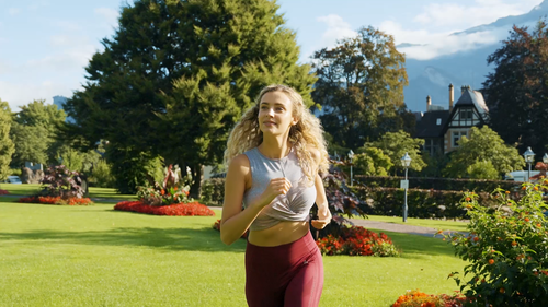 Eine Frau joggt unter einem strahlend blauen Himmel durch einen farbenfrohen Park. Sie trägt ein graues Tanktop und kastanienbraune Leggings und lächelt vor dem Hintergrund von Bäumen und Blumen.