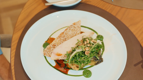 Ein Gourmetgericht mit geschnittenem Hähnchenfleisch und einem mit Sesam bestreuten Cracker auf einem Bett aus Blattgemüse und Nüssen, kunstvoll garniert mit grüner und roter Sauce.