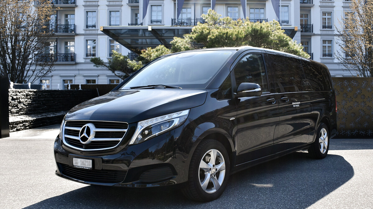 Ein schwarzer Mercedes-Benz-Van steht vor einem eleganten Gebäude mit Balkonen. Sonnenbeschienene Bäume umgeben das Gelände und vermitteln einen luxuriösen und raffinierten Eindruck.