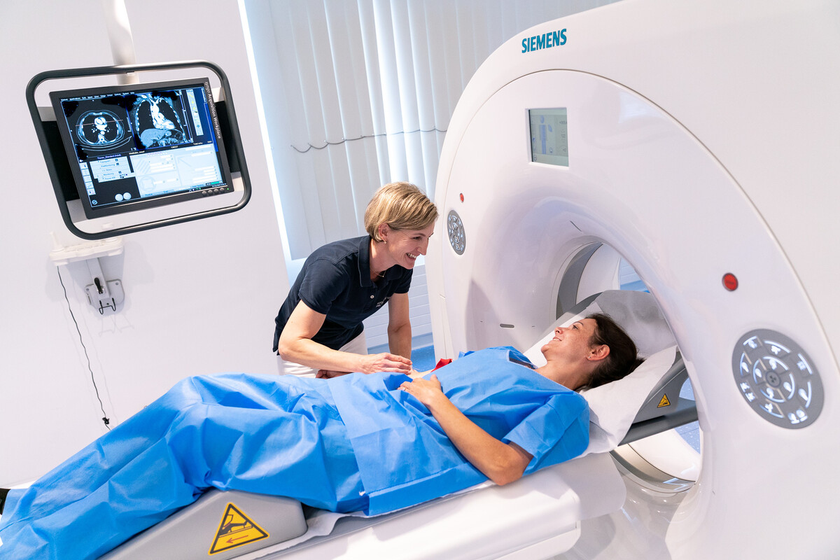 Ein medizinisch-technischer Assistent hilft einem Patienten in einem blauen Kittel auf dem Bett eines CT-Scanners. Auf einem Bildschirm werden die Scanbilder angezeigt. Die Szene vermittelt eine beruhigende Atmosphäre.