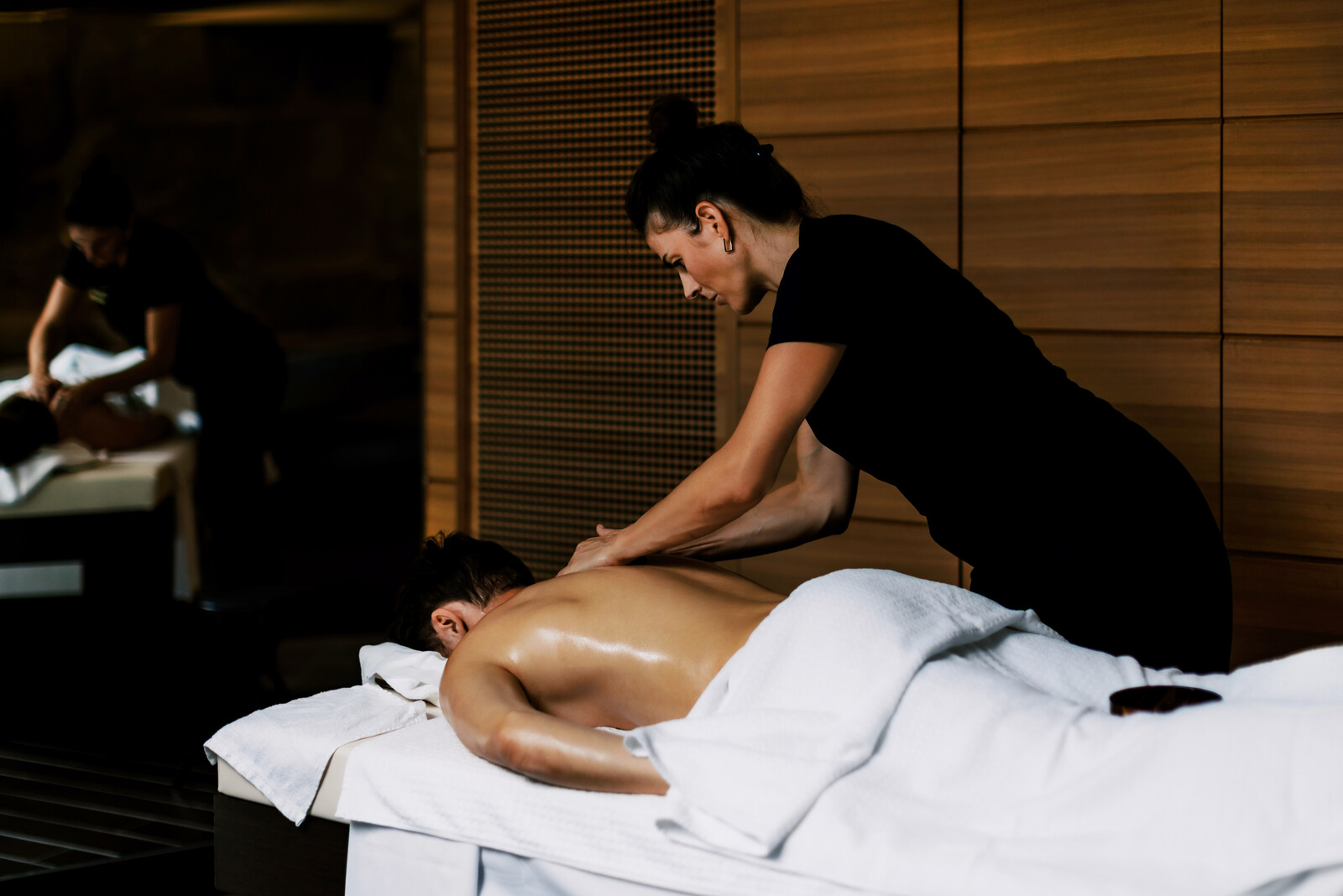 A massage therapist in a black outfit gives a back massage to a client lying on a table, with a calm, relaxing spa ambiance. A mirror reflects the scene.