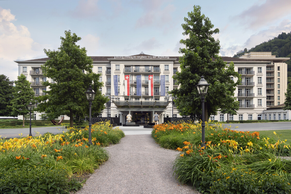 The hotel entrance to the Grand Resort Bad Ragaz with colorful flower gardens and trees lining a gravel path. Swiss and blue flags hang above the entrance, exuding elegance.