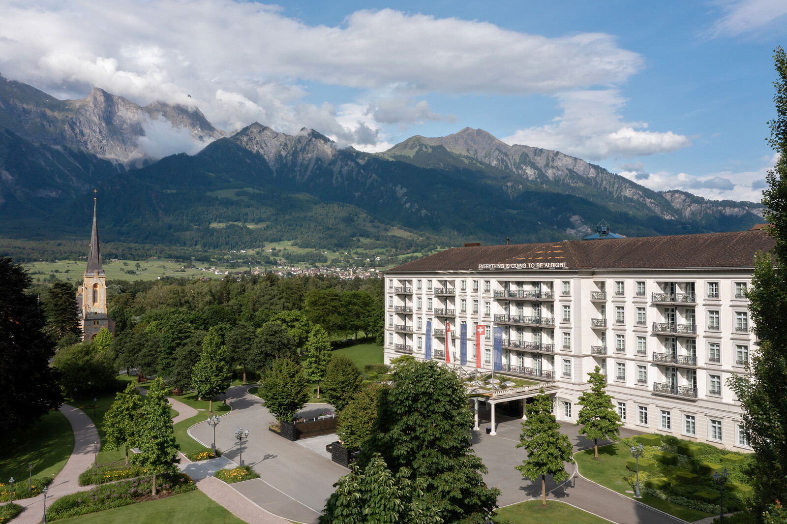 The Grand Hotel Bad Ragaz with flags, surrounded by lush greenery and a picturesque mountain backdrop. Church tower visible. The sky is partly cloudy, serene ambience.