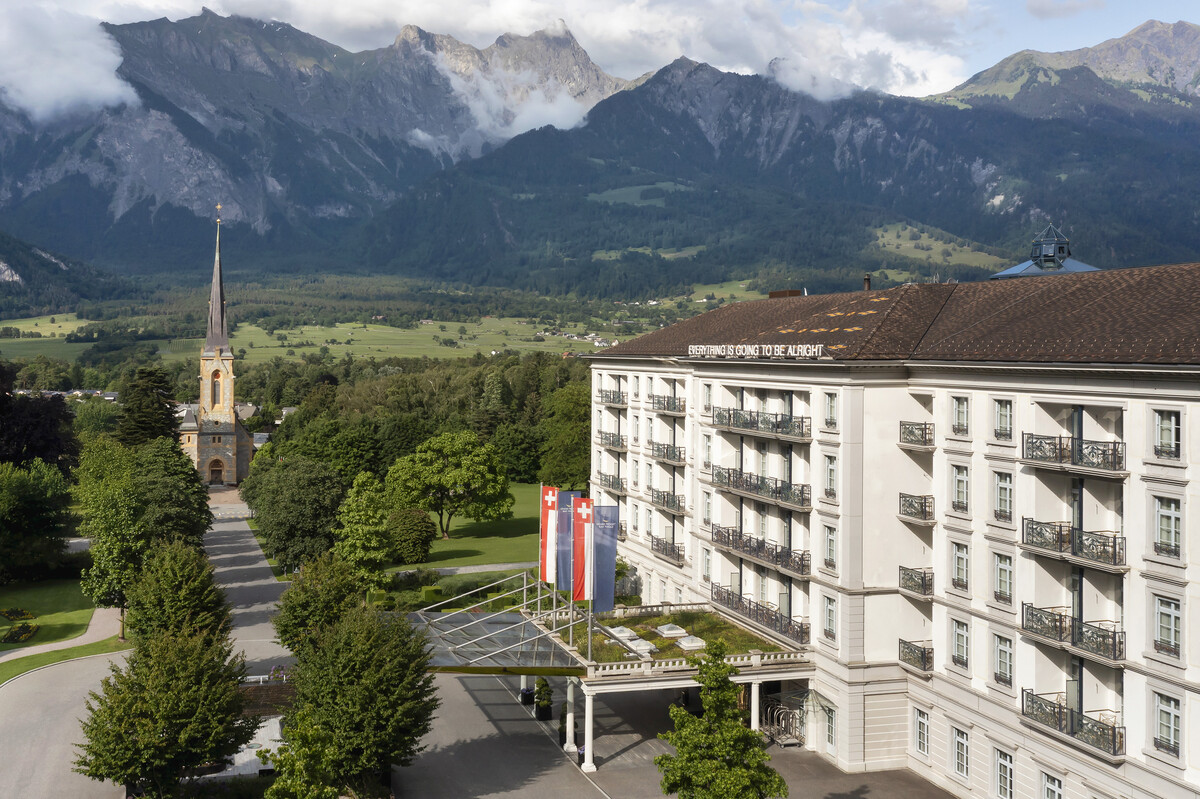 The Grand Resort Bad Ragaz with its Swiss flags is set against a backdrop of lush greenery and majestic mountains. A tall church tower stands on a tree-lined path.