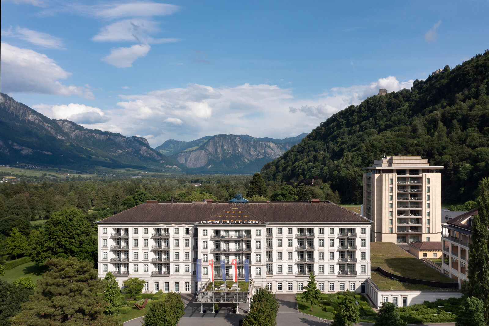 Grand Hotel Bad Ragaz mit weisser Fassade und Balkonen, flankiert von üppigem Grün und Bergen. Fahnen und „Alles wird gut“-Schilder geben Hoffnung.
