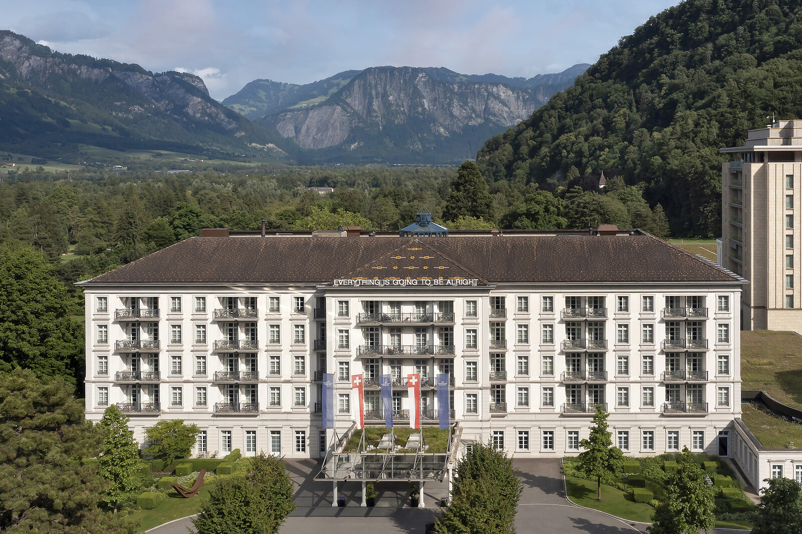 Grand Hotel Bad Ragaz mit weißer Fassade und Balkonen, flankiert von üppigem Grün und Bergen. Fahnen und „Alles wird gut“-Schilder geben Hoffnung.
