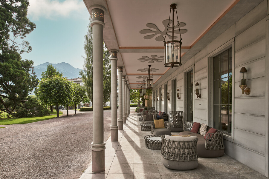 The hotel's covered arcade running the length of the facade, slim columns opening onto the gravel drive and parkland on one side and woven armchairs set along the wall on the other.