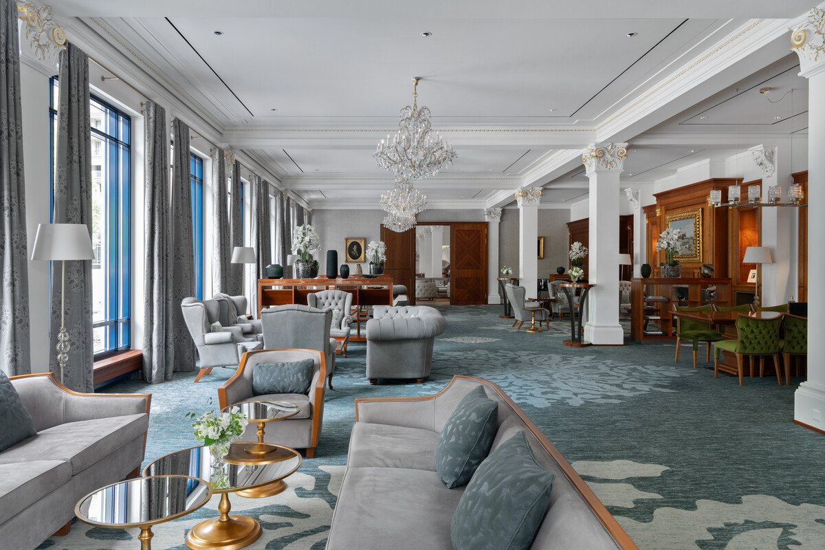 Der grüne Salon des Grand Resort Bad Ragaz mit eleganten grauen Sofas, goldenen Akzenttischen und Plüschsesseln auf einem gemusterten blauen Teppich. Kristallkronleuchter hängen von der Decke und schaffen eine elegante Atmosphäre.