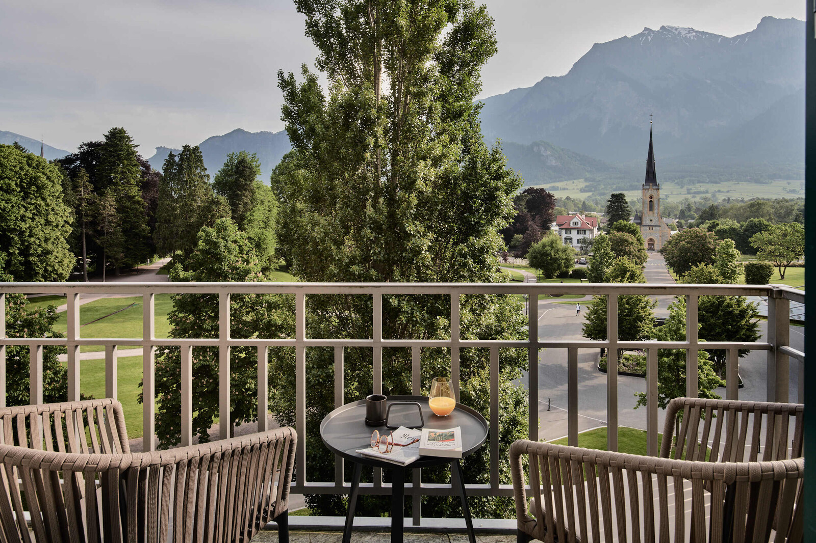 Blick vom Balkon eines Comfort Zimmers im Grand Hotel Hof Ragaz. Auf dem Balkon befinden sich zwei grosszügige Sessel und ein Tischchen mit Platz für Getränke, Bücher. Im Hintergrund ist Bad Ragaz und die traumhafte Bergkulisse zu sehen.