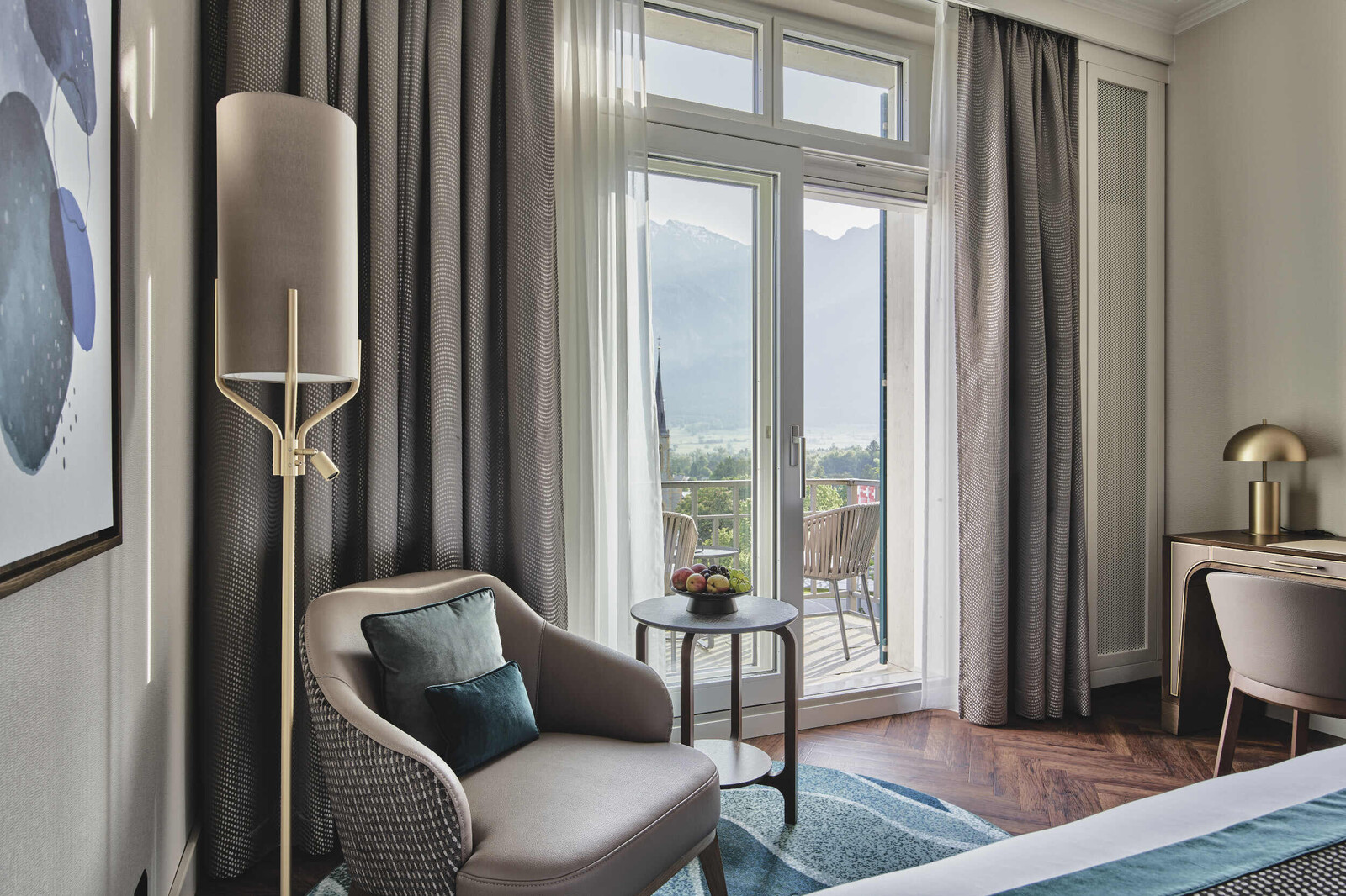 Blick schräg vom Fusse des Betts in Richtung der Fenster. Links vom Fenster befindet sich ein Sessel mit Beistelltisch und Stehlampe. Rechts vom Fenster steht ein Schreibtisch mit Stuhl.