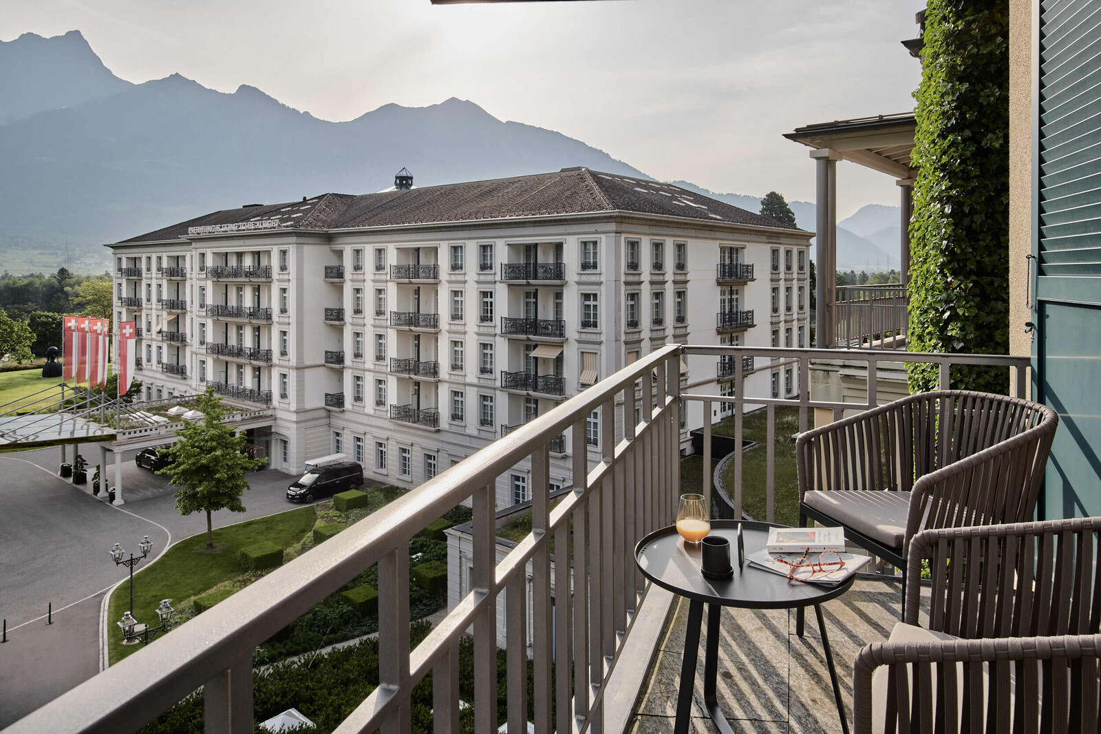 View from the corner of a balcony in the direction of Grand Hotel Quellenhof. On the balcony are two armchairs and a table. In the background you can see a mountain landscape.