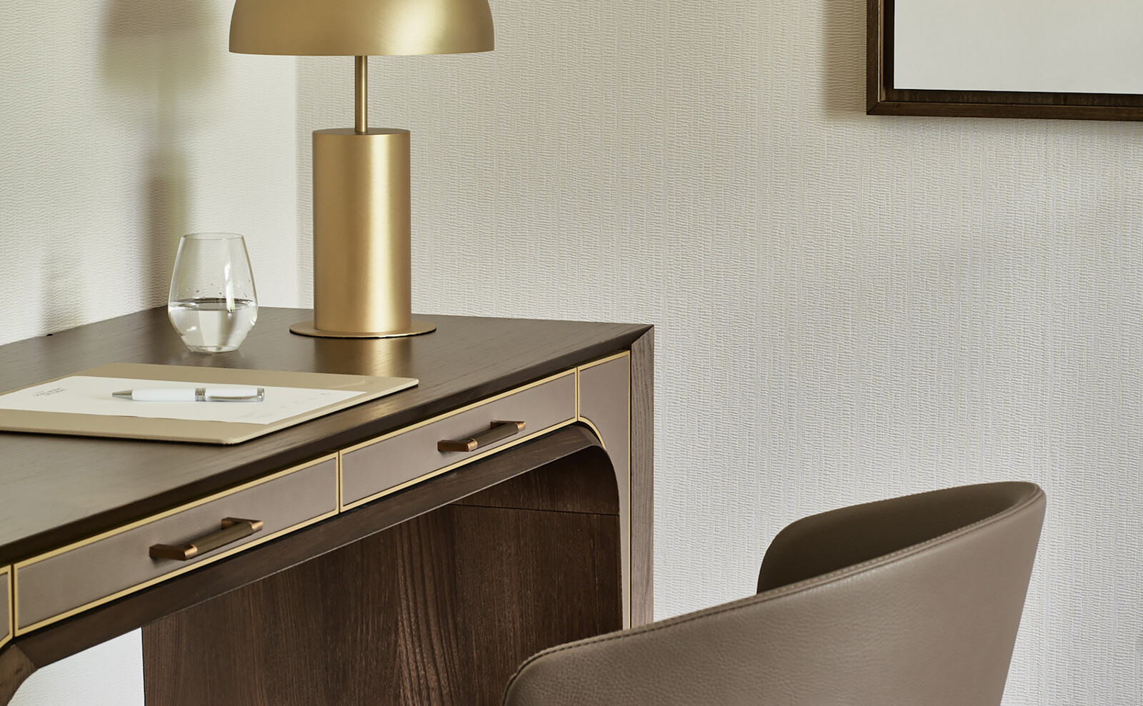 Nahaufnahme mit schrägem Blick auf einen Schreibtisch vor dem ein brauner Stuhl mit Lederarmlehne steht. Auf dem Tisch befindet sich ein Glas Wasser, eine Tischlampe sowie ein Papier mit Stift.