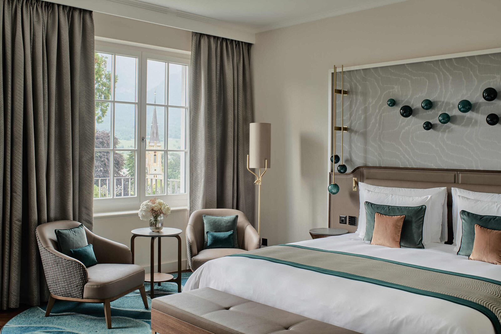 View diagonally past the bed to the window of a Grand Deluxe Room in the Grand Hotel Hof Ragaz. In front of the window are two armchairs and a side table.