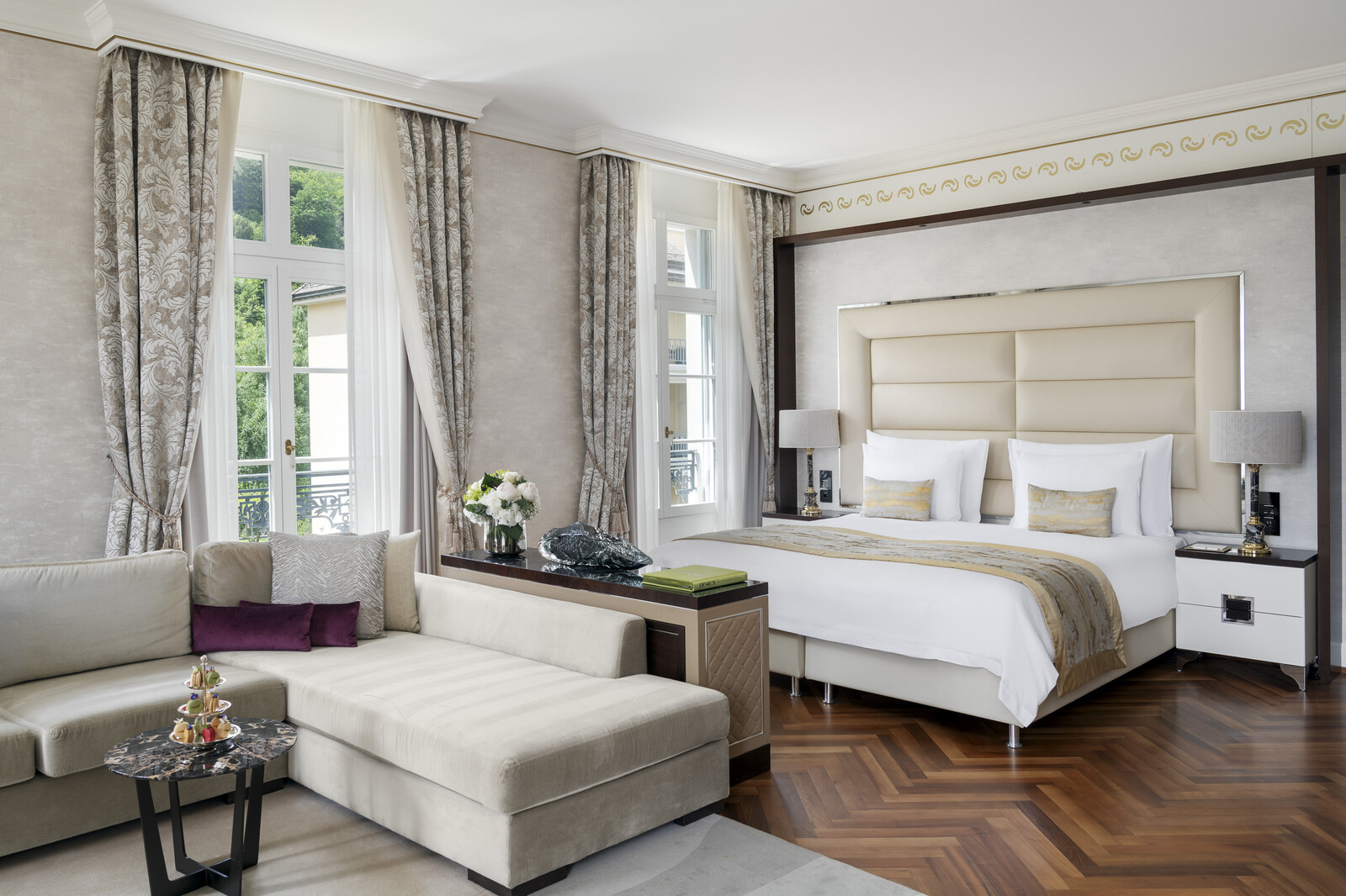 Die luxuriös eingerichtete Junior Eck Suite im Quellenhof mit Fischgrätenparkett, zwei grossen Fensterfronten, einem Queensize Bett und einer gemütlichen Sofaecke in den Farben Gold und Beige.