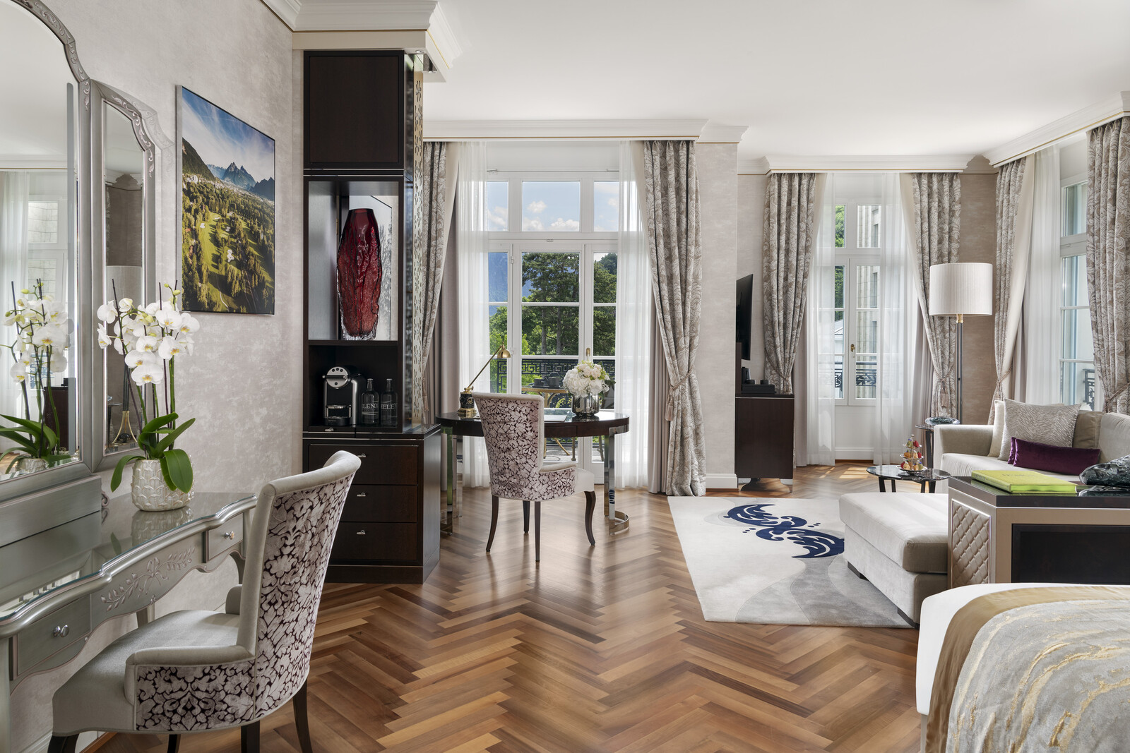 Elegant, spacious living room with herringbone wood floors, light gray walls, large windows, chic furniture, and decor. Bright, airy, and inviting atmosphere.