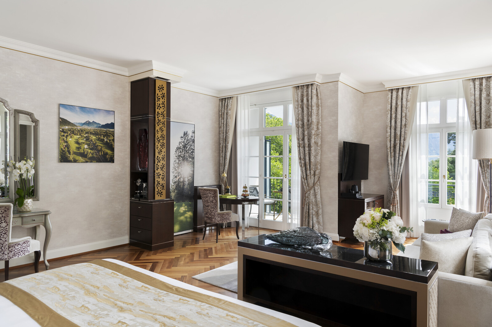 The luxuriously furnished Junior Corner Suite at Quellenhof in the colors of gold and beige with herringbone parquet flooring, two large window fronts, a comfortable sofa corner, a TV, a desk and chair.