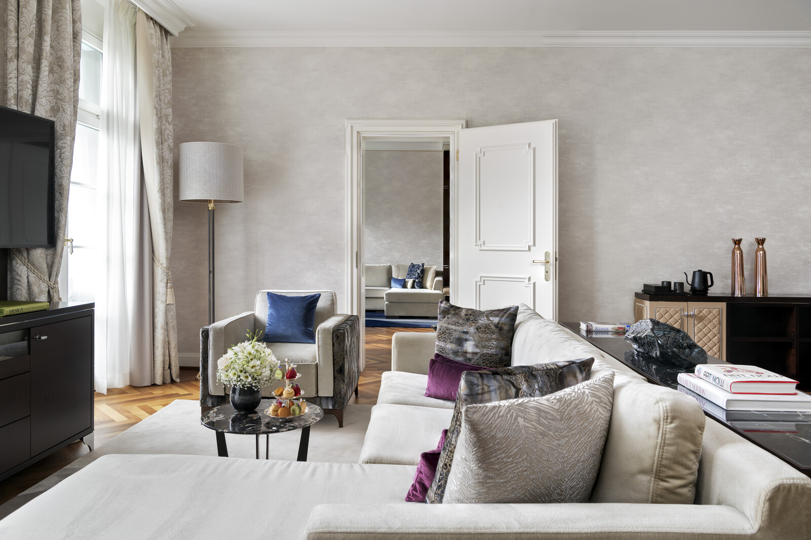 Elegant living room with neutral tones, featuring a plush sofa, colorful cushions, a small round table with flowers, and a large open door. Bright and inviting ambiance.