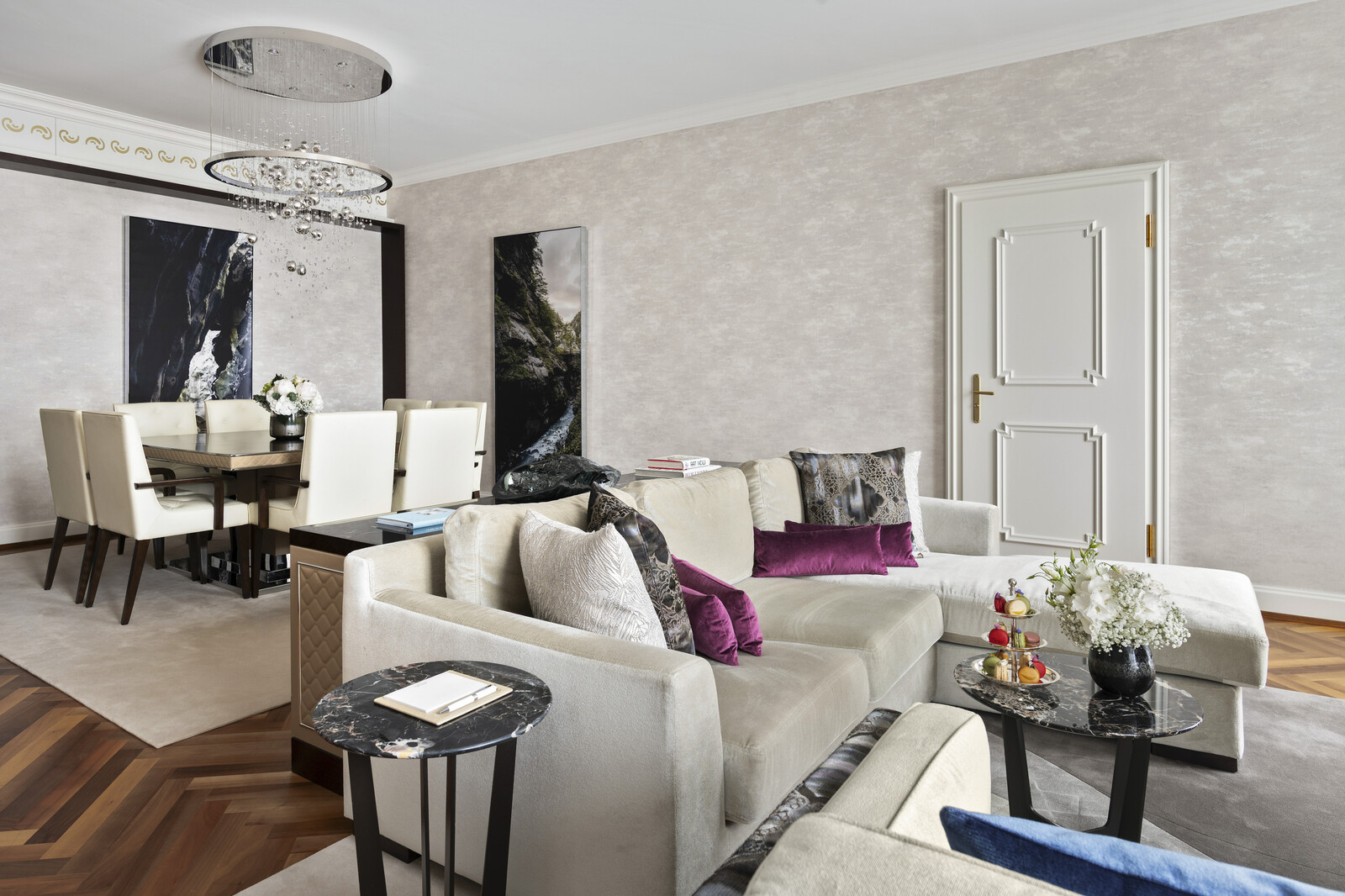 Elegant living room with a cream sofa, purple cushions, and abstract art. Dining area with white chairs. Modern, refined atmosphere.