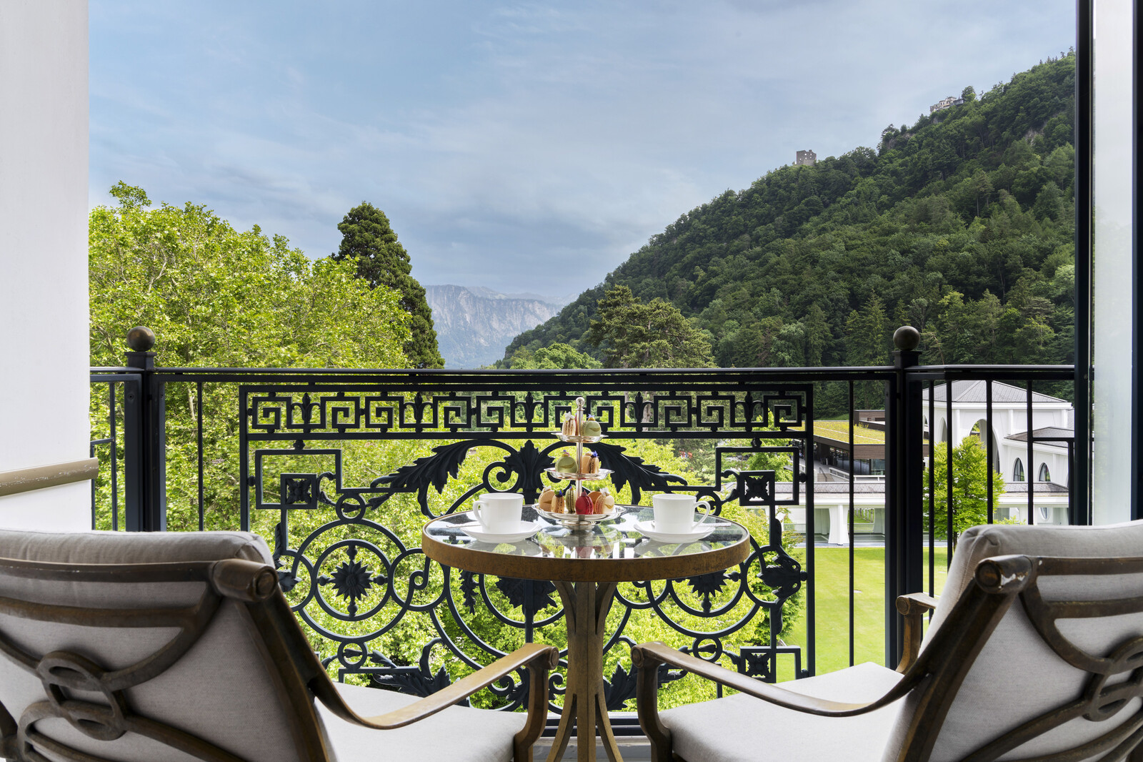 Ein kleiner runder Tisch mit Blumen und Teetassen steht auf einem Balkon. Ein verziertes Geländer bietet einen Blick auf üppig grüne Hügel unter blauem Himmel und vermittelt Ruhe.