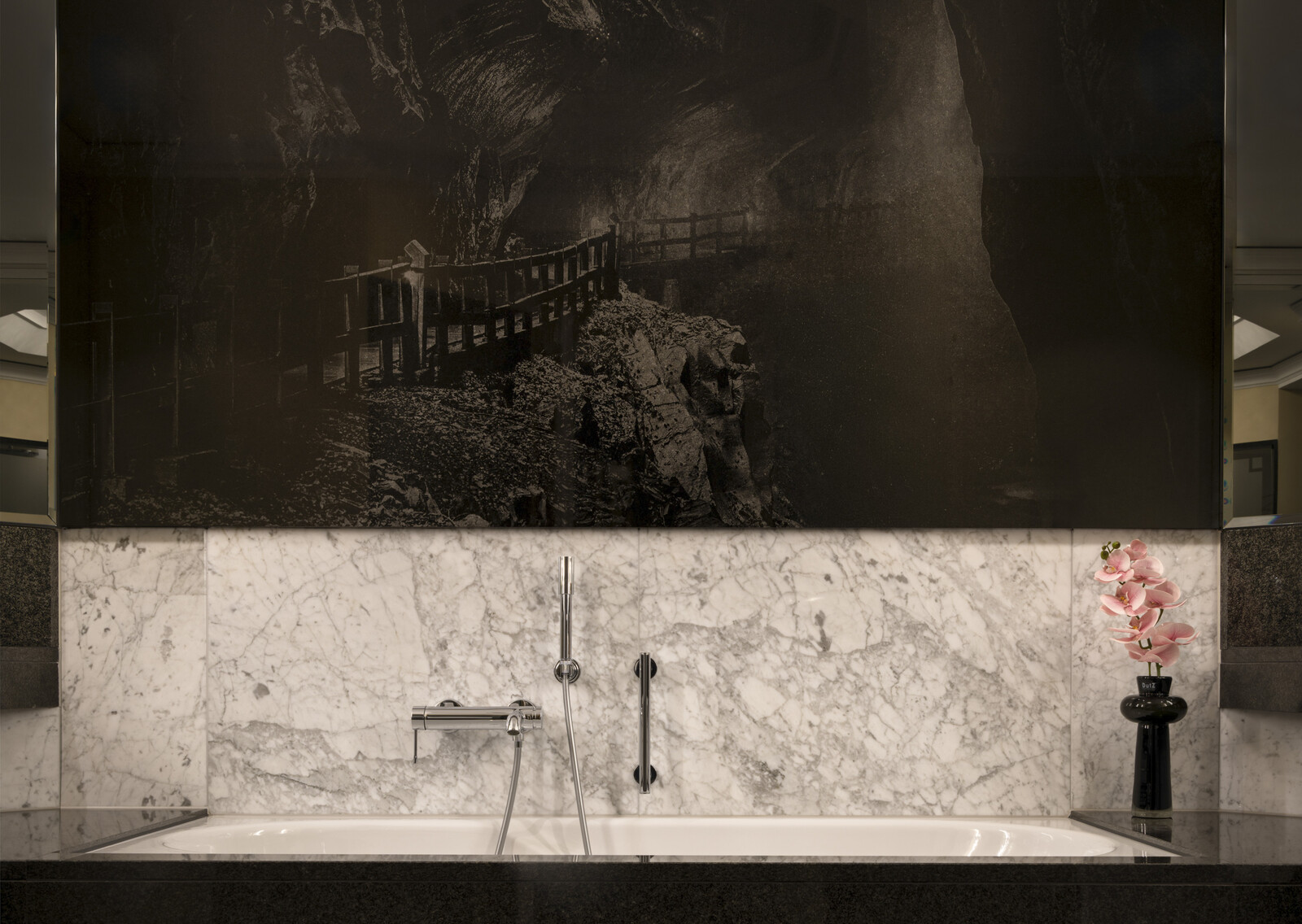 Luxuriöses Badezimmer mit weisser Marmorbadewanne, Akzenten aus poliertem schwarzem Stein und einer auffälligen schwarz-weissen Wandmalerei in den Bergen. Elegante und heitere Atmosphäre.