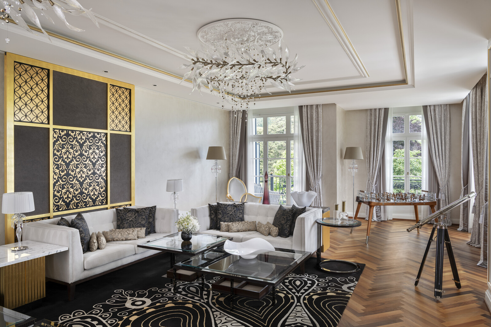 Elegant living room with luxurious decor features a white sofa, ornate gold and black patterned wall, crystal chandelier, and large windows with sheer curtains.