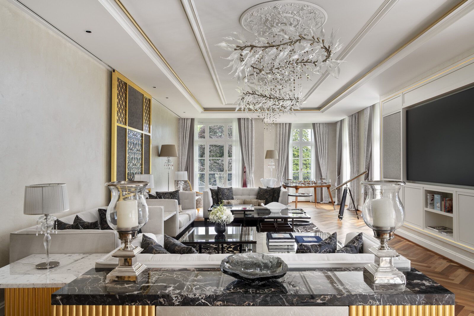 Elegant living room with ornate chandeliers, large TV, and plush seating. Neutral tones, wooden floor, telescope by window, and modern decor create a luxurious feel.