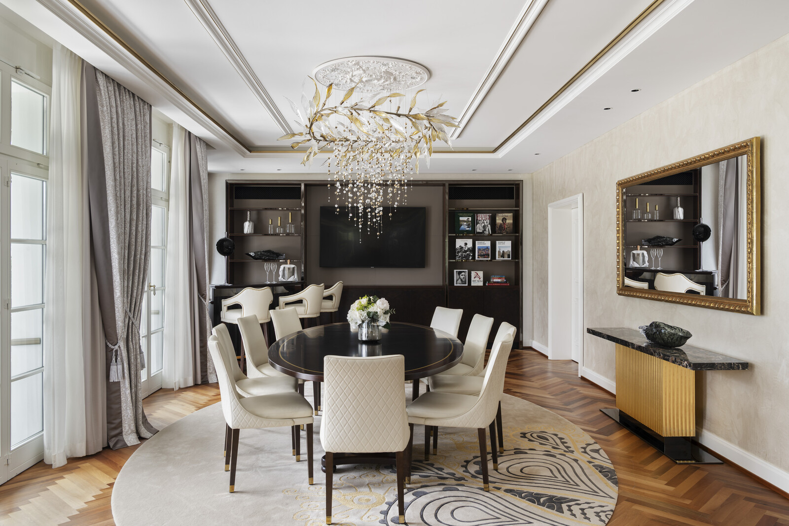 Elegant dining room with a large round table, cream chairs, and a floral centerpiece. A golden chandelier hangs above.