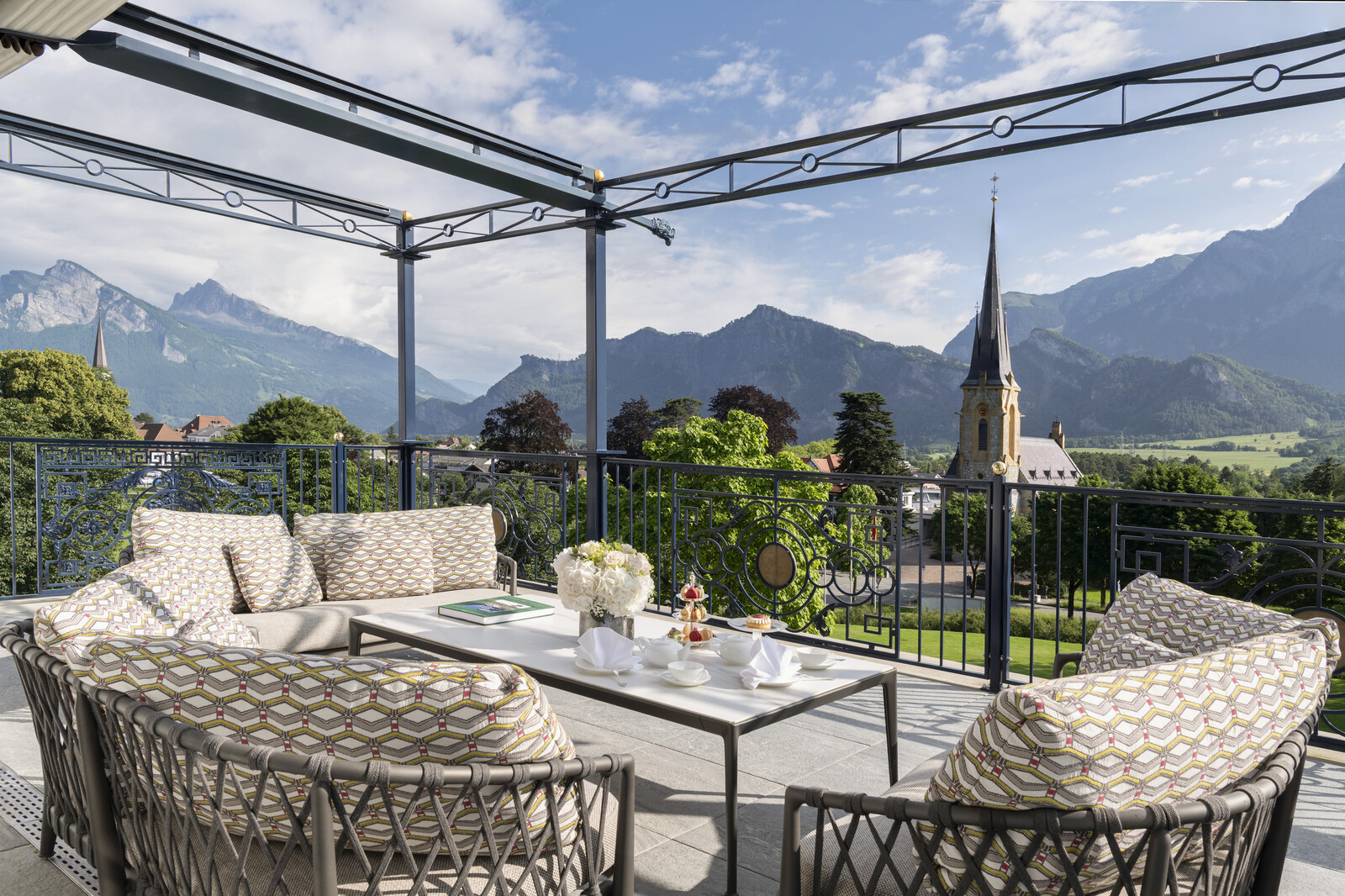 Cozy outdoor seating with patterned cushions on a balcony overlooking majestic mountains and a church steeple.
