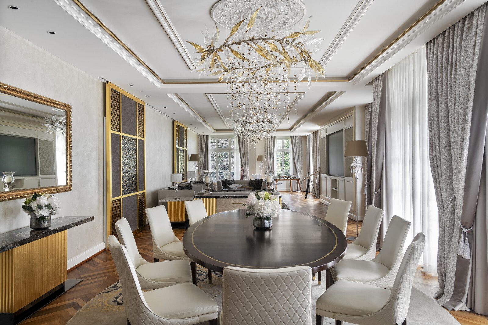 Elegant dining room with a round dark table, white cushioned chairs, ornate chandelier, large windows with drapes, and a floral centerpiece.