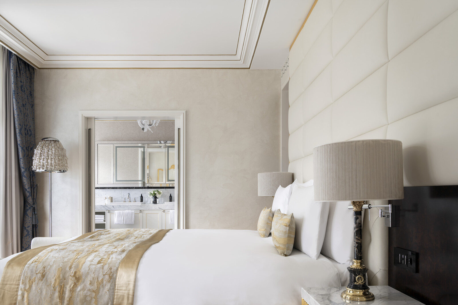 Elegant hotel room with neutral tones, featuring a plush bed and pillows, a floor lamp, a TV, and an open view into a stylish bathroom.