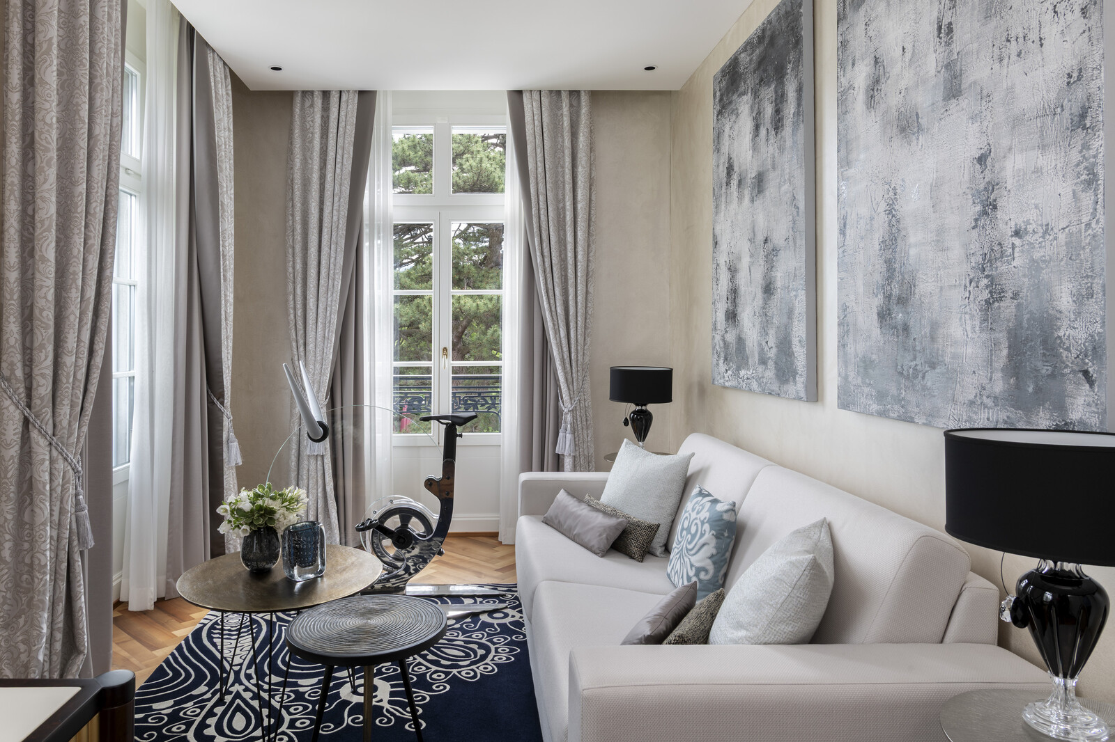 Elegant room with a neutral-toned sofa, textured cushions, abstract paintings, a round coffee table, and a stationary bike near sheer drapes and a tall window.