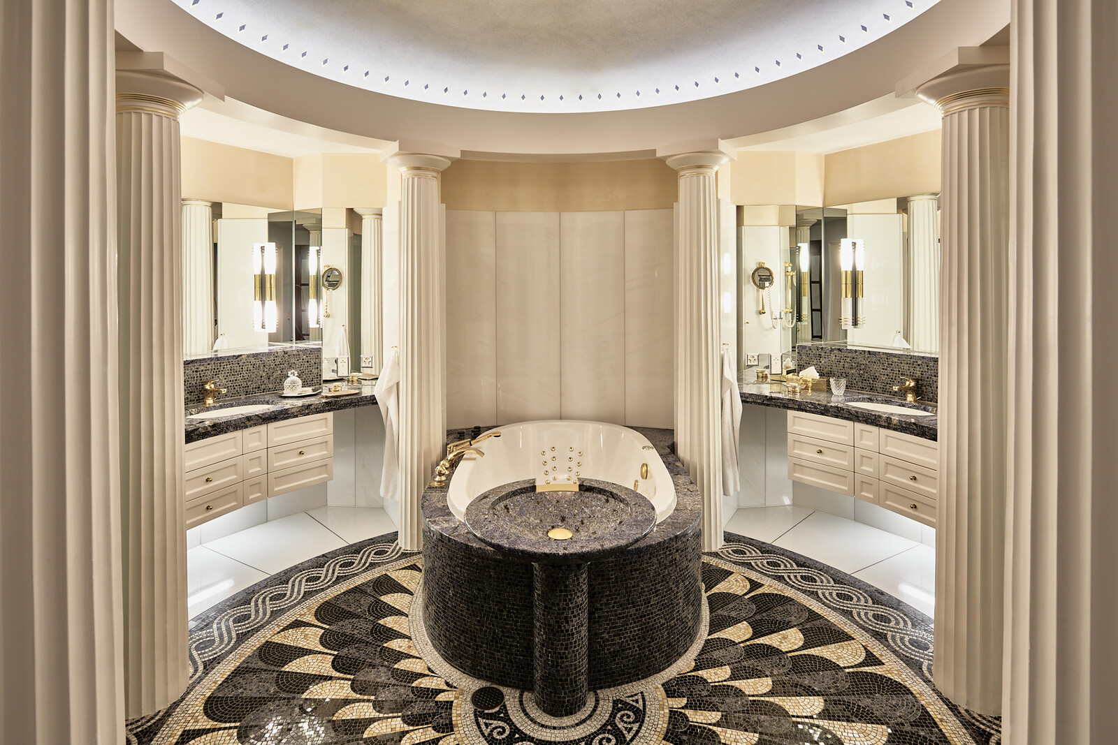 Luxurious bathroom with a central circular tub, ornate columns, and intricate black and gold flooring. Mirrors and soft lighting create an opulent ambiance.