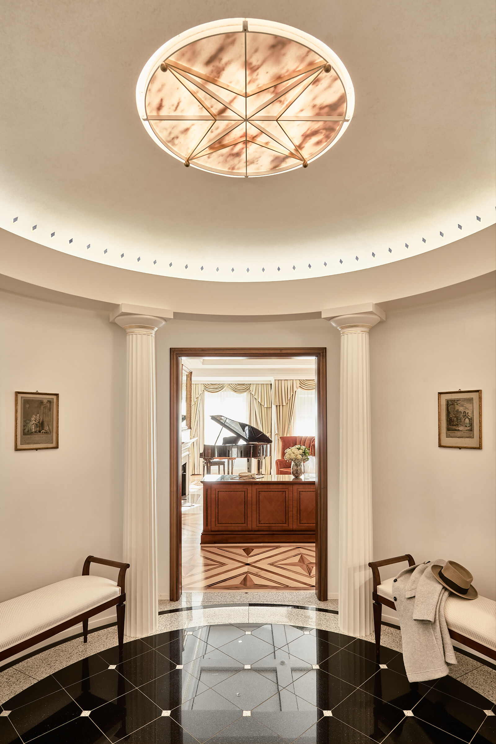 Elegant foyer with a circular ceiling light, white walls, bench seating, and a view into a room with a grand piano.