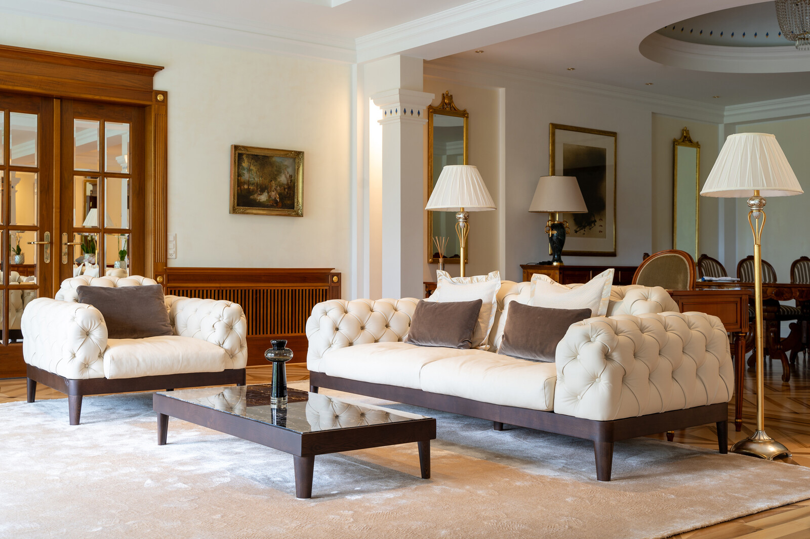 Elegant living room with a cream sofa and armchair, dark wood accents, and floor lamps. Dining area in the background, exuding warmth.