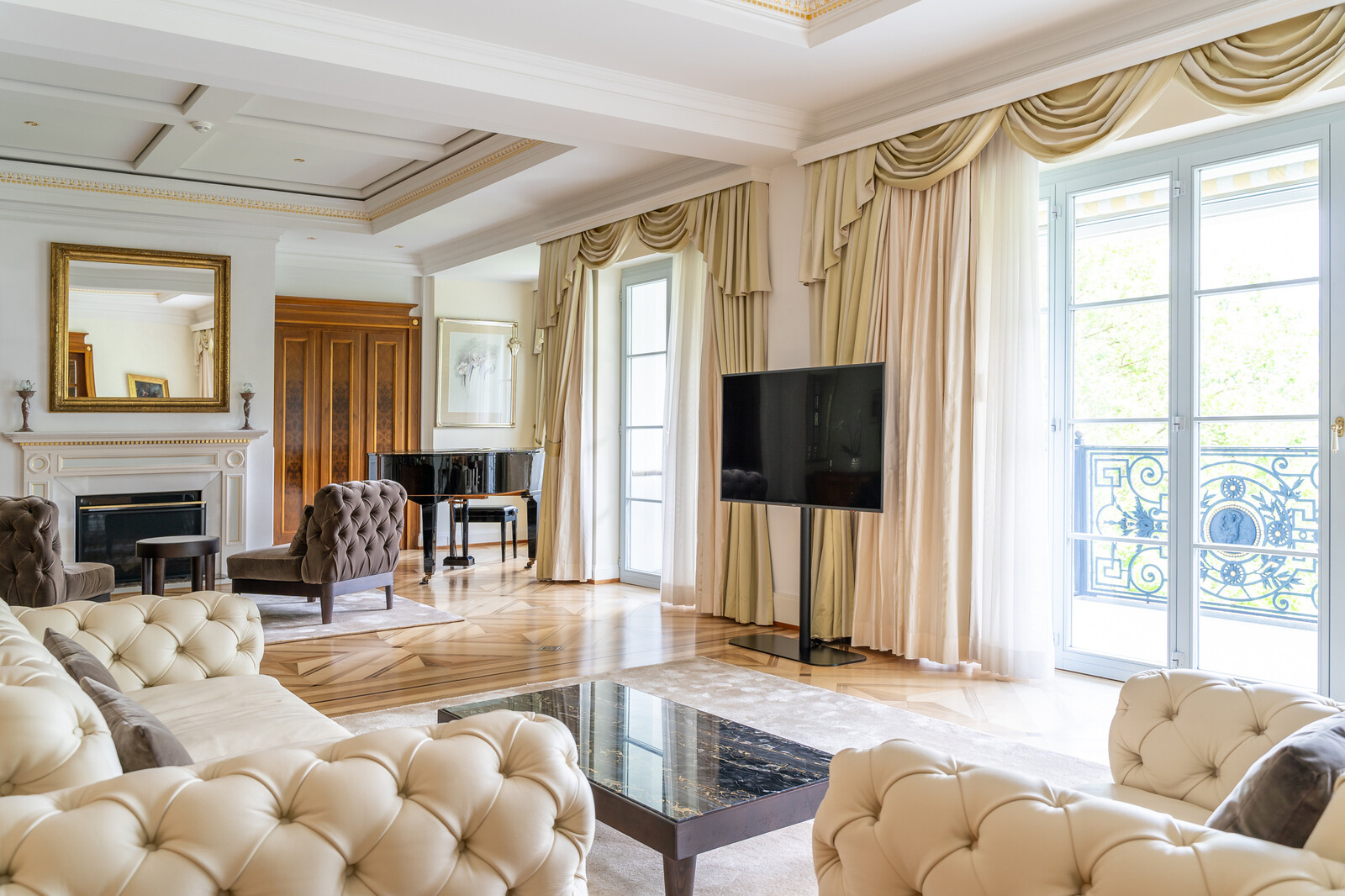 Elegant living room with beige tufted sofas, glass coffee table, and large windows with curtains. A piano and fireplace add a refined touch.