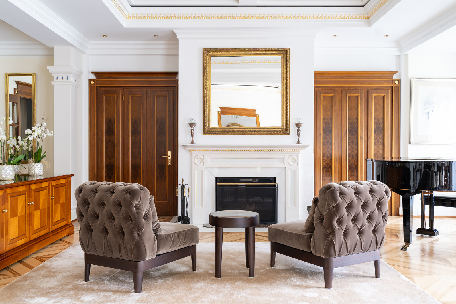 Elegant living room with two tufted armchairs facing a small table, set before a white fireplace. A mirror hangs above, with a grand piano and wooden doors nearby.