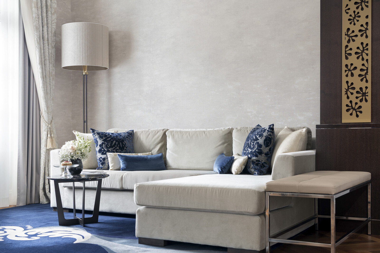 Anspruchsvolles Wohnzimmer mit einem beigen Sektionssofa mit blauen Kissen, einem Couchtisch aus dunklem Holz, einer modernen Stehlampe und einem blauen Plüschteppich. Gemütliches Ambiente.