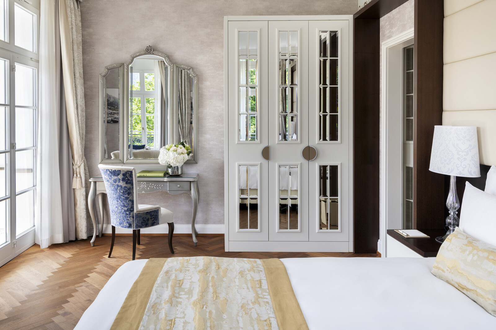 Elegantes Schlafzimmer mit grossem verspiegeltem Kleiderschrank, gemütlichem Bett mit goldfarbener Bettwäsche, verziertem Waschtisch mit frischen Blumen und sanftem Tageslicht.