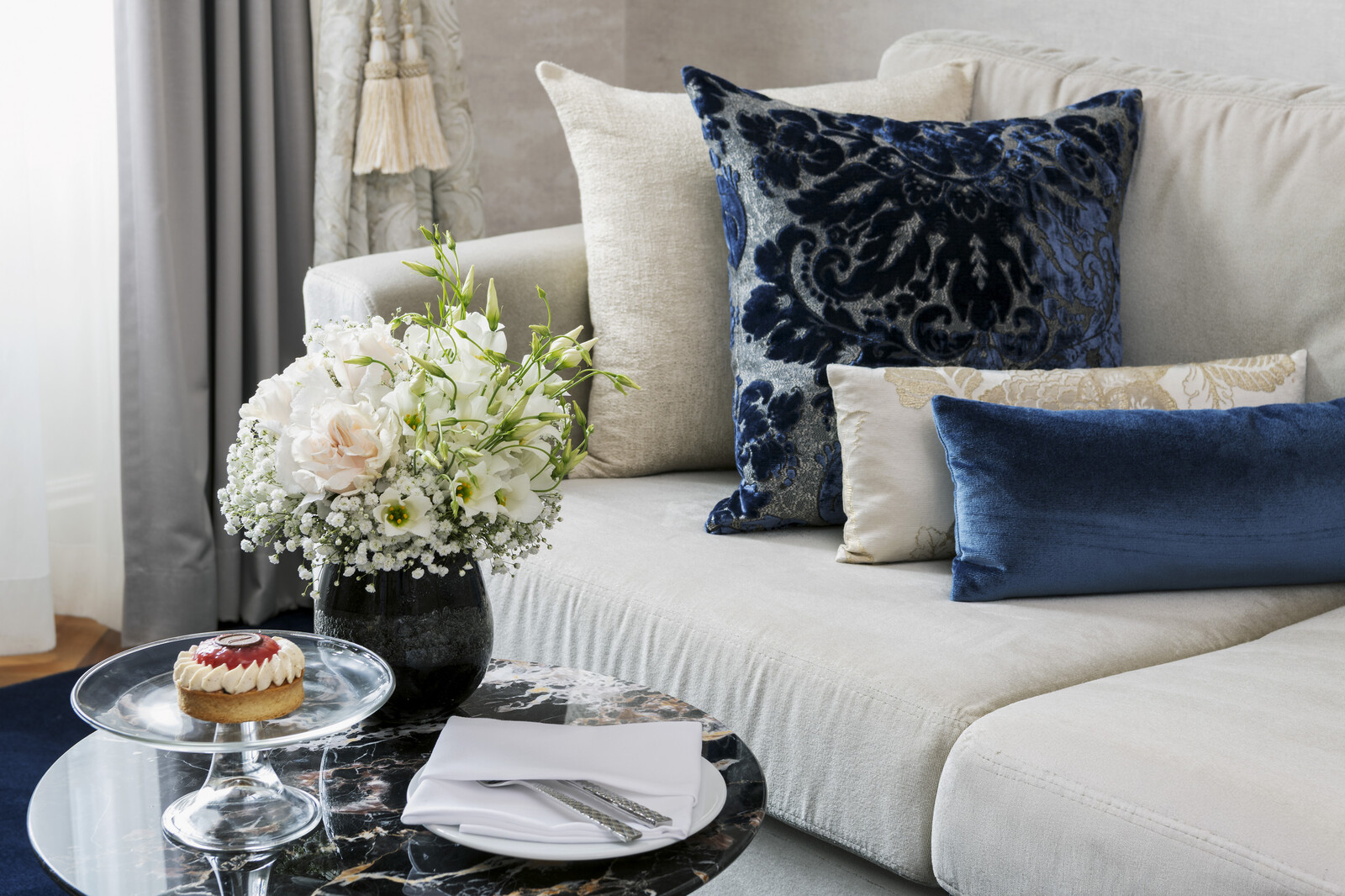 Elegantes Wohnzimmer mit einem beigen Sofa, das mit plüschigen blauen und gemusterten Polstern geschmückt ist. Auf einem Marmortisch stehen ein Blumenstrauß, ein Dessert und gefaltete Servietten, die eine gemütliche, einladende Atmosphäre schaffen.