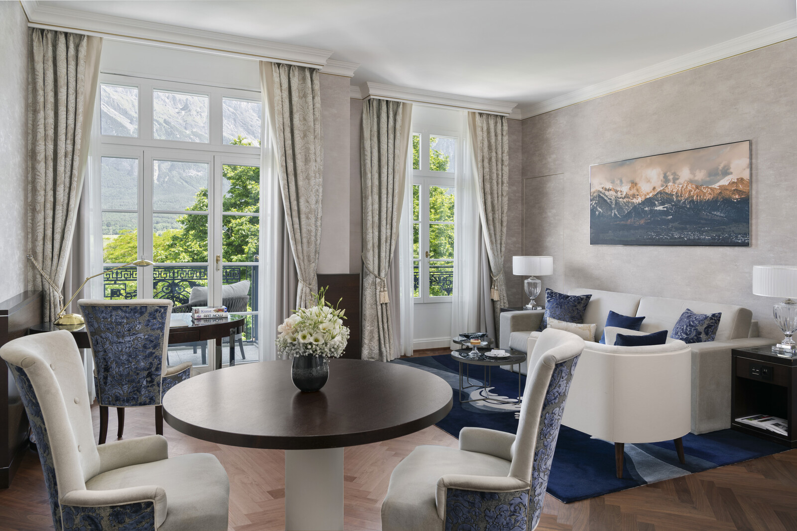 Elegantes Wohnzimmer mit großen Fenstern, Bergblick und neutralen Tönen. Mit Plüschsesseln, rundem Tisch und Landschaftsgemälde. Helles und heiteres Ambiente.