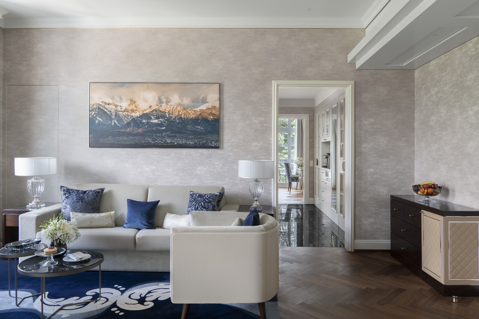 Elegantes Wohnzimmer mit einem beigen Sofa, blauen Kissen und einem Gemälde mit einer Berglandschaft. Auf einem gläsernen Couchtisch stehen Blumen, die ein ruhiges Ambiente vermitteln.