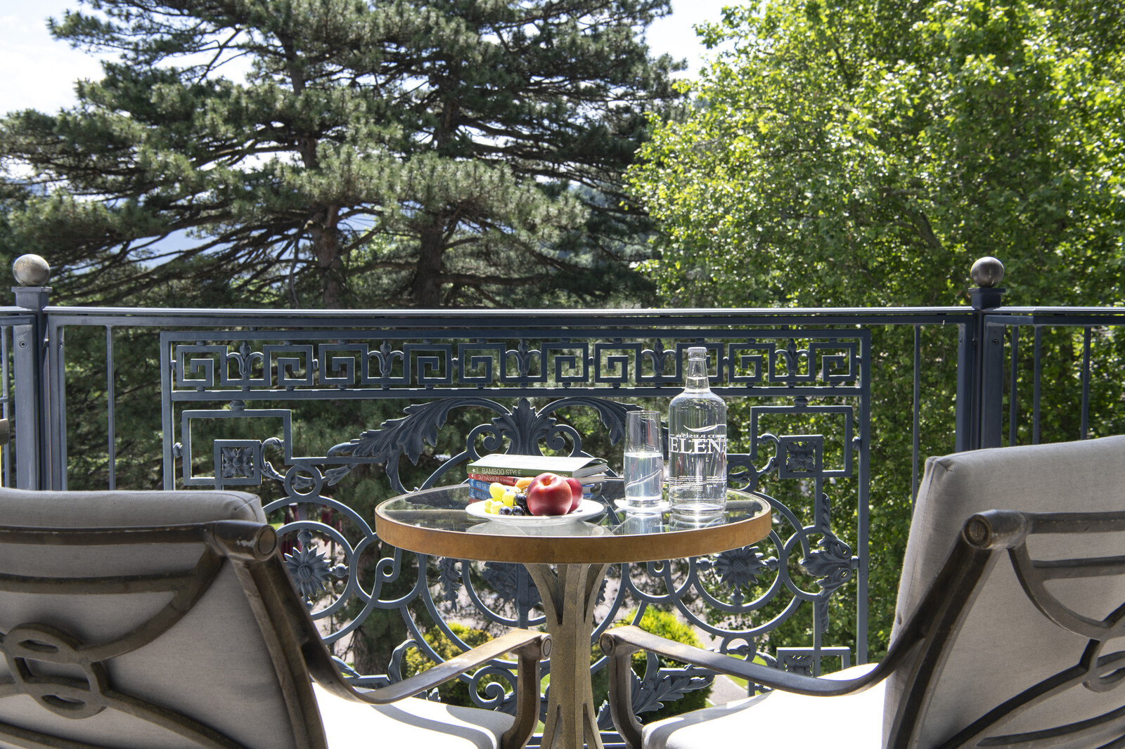 Ein ruhiger Balkon mit zwei Stühlen vor einem kleinen runden Tisch mit einer Wasserflasche und einem Teller mit Äpfeln, eingerahmt von üppigen grünen Bäumen im Hintergrund.