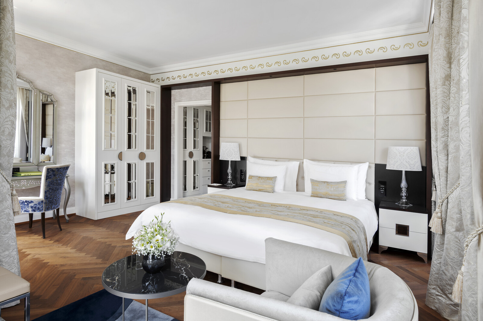 Luxurious hotel room with a large bed, elegant white and gold decor, plush armchair, blue and gold accents, and a vase of flowers, conveying comfort and sophistication.