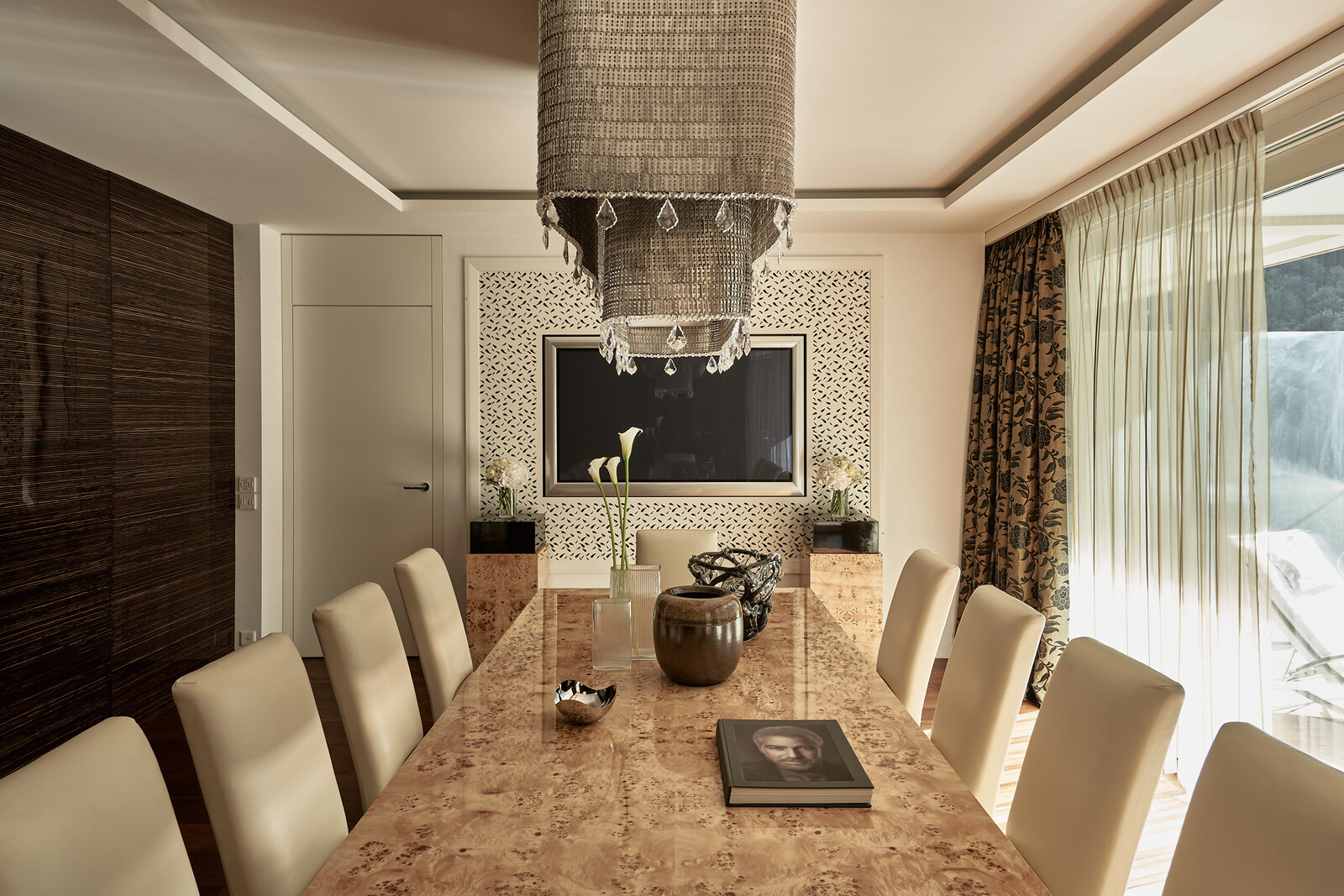 Elegantes Esszimmer mit einem langen Marmortisch und beigen Stühlen. Darüber hängt ein großer Kronleuchter. Ein Buch und eine Vase stehen mittig auf dem Tisch.