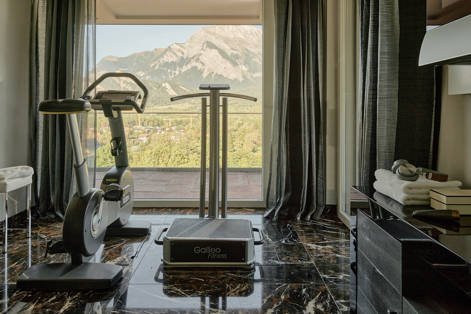 Elegantes Fitnessstudio mit einem stationären Fahrrad und einem Laufband auf dunklem Marmorboden.Ein großes Fenster bietet einen atemberaubenden Blick auf die Berge.