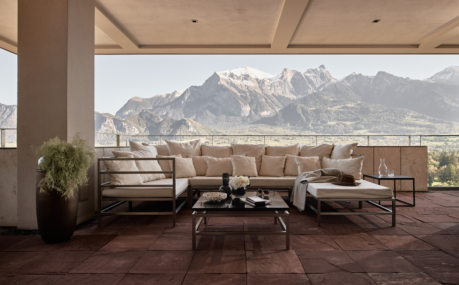 Geräumige Terrasse mit einem modernen beigen Sektionssofa mit Blick auf schneebedeckte Berge. Die minimalistische Einrichtung umfasst einen zentralen Tisch mit Blumen.