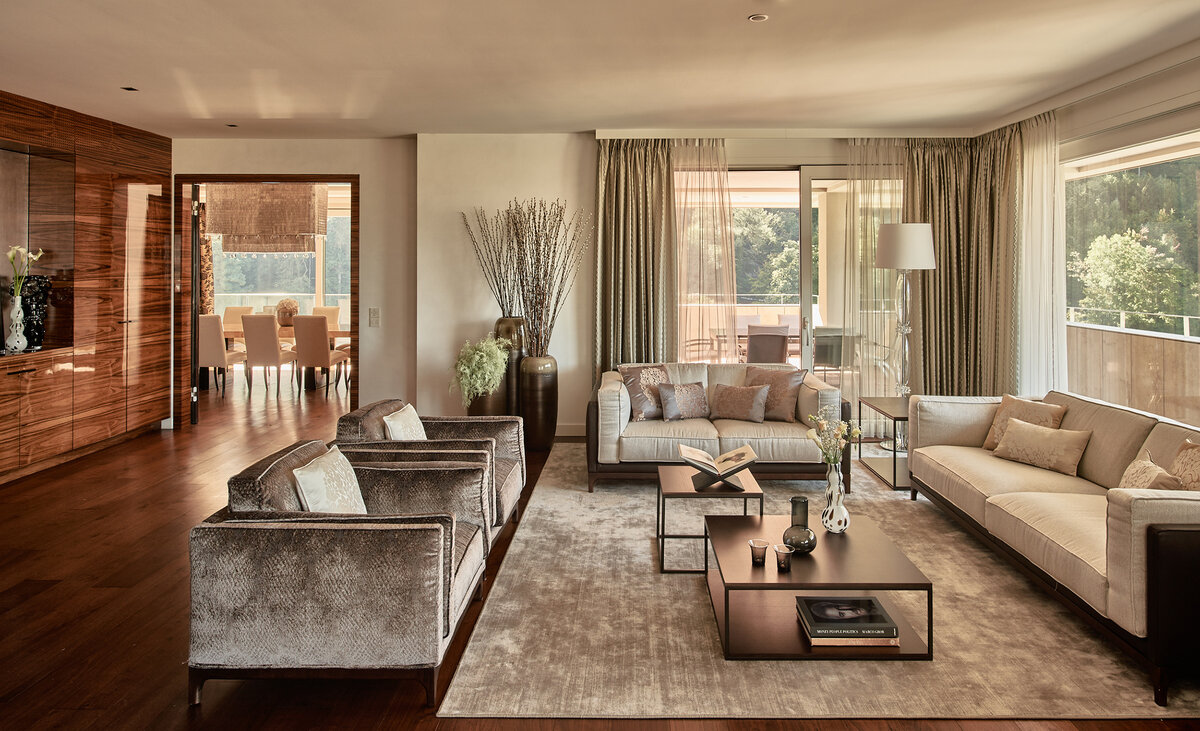 Ein modern eingerichtetes Wohnzimmer mit grossen Fensterfronten, beigen Wohnsesseln und passender Dekoration im Penthouse im Grand Resort Bad Ragaz.